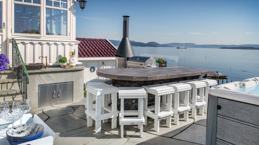 En terrasse ved sjøen med bruddheller på bakken og et utekjøkken med en stor bar og boblebad.