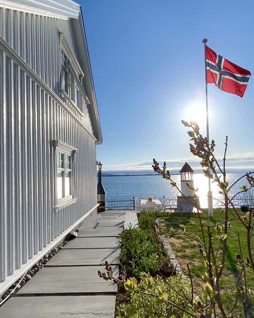 En gångstig med trampsten av skiffer som går längs med ett hus och leder ner till vattnet. En norsk flagga vajar i vinden i trädgården.