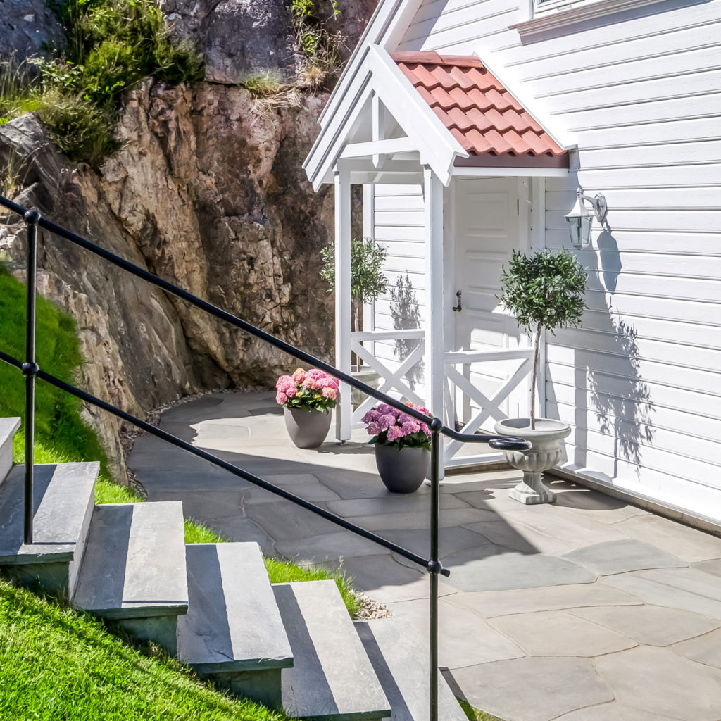 A staircase with slate steps going up from a terrace paved with crazy paving outside a white cottage.