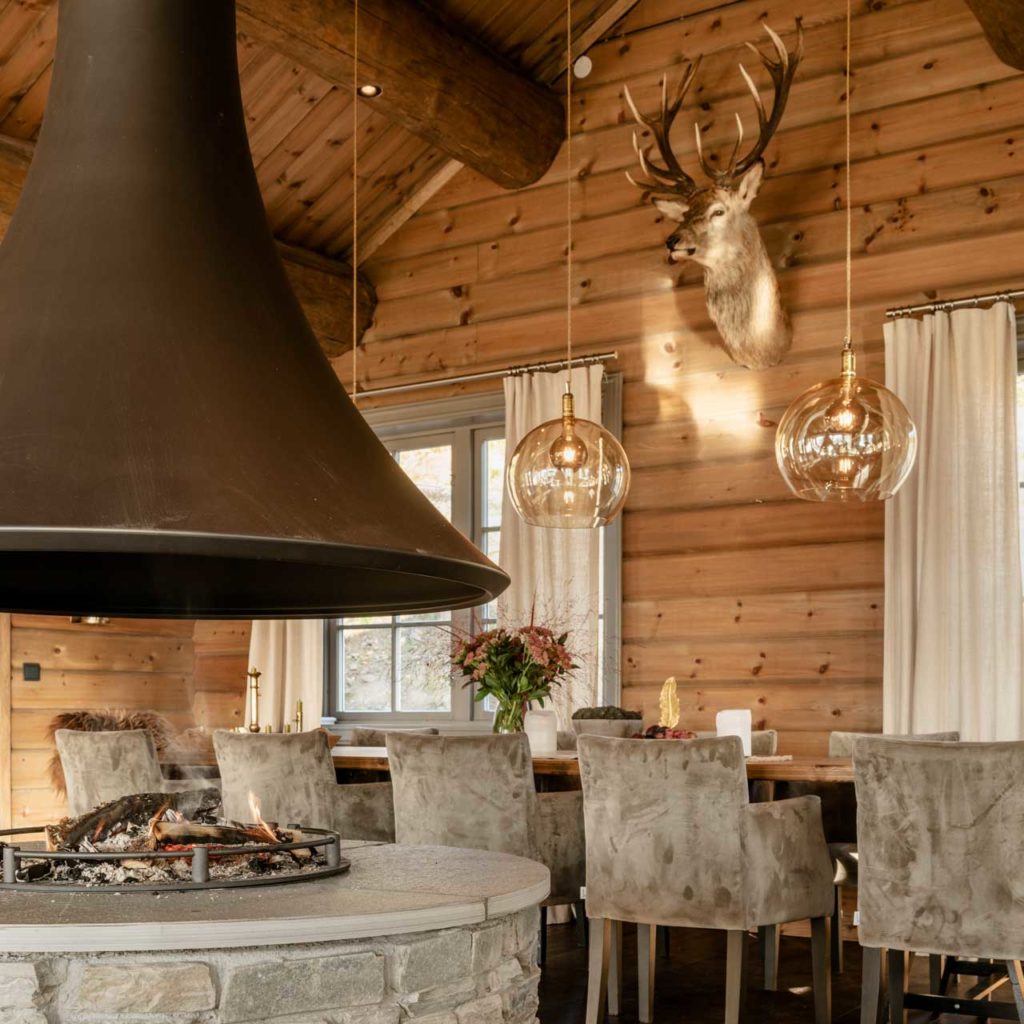 Round fireplace built of slate bricks. In the background is a dining table and on the wall there is a stuffed reindeer.