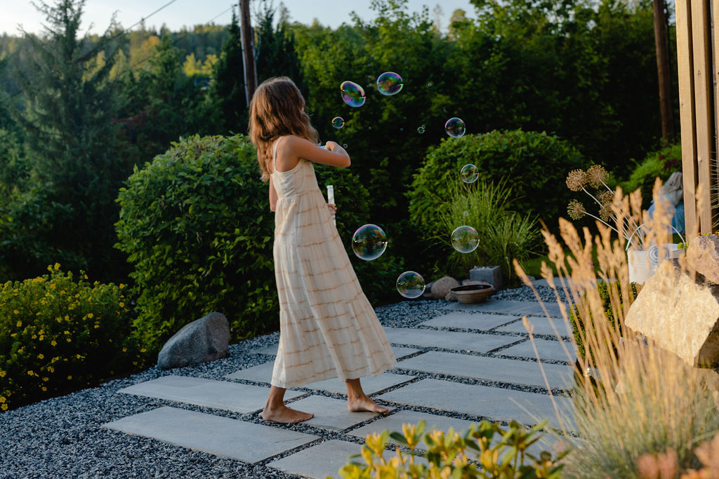 A girl in a summer dress blowing soap bubbles while walking barefoot on slate slabs laid on gravel in a driveway.