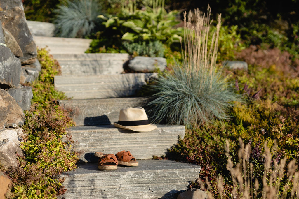 A slate garden natural stone staircase on a slope with beautiful plants on the sides. A hat and a pair of shoes lie on each slate step.