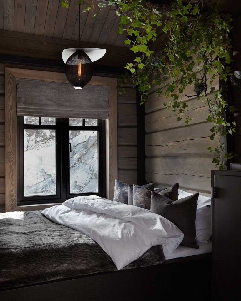 Et mørkt og luksuriøst soverom på en hytte med tømmervegger, grønne plante og vakkert oppredd seng med flotte tekstiler