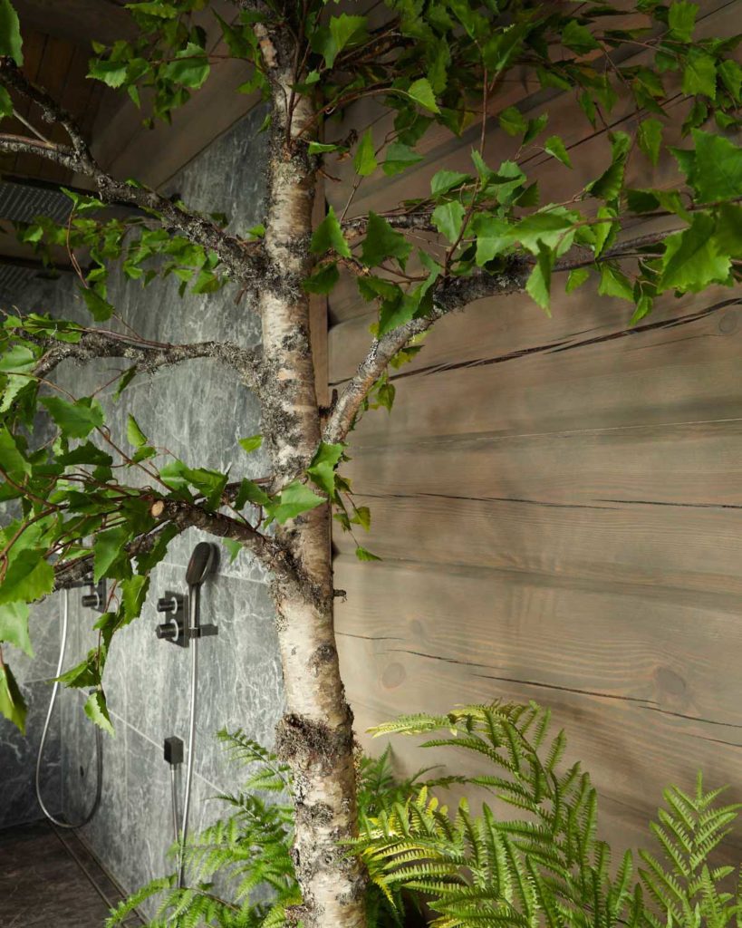 Et bad med fliser av naturstein i kombinasjon med tømmervegger. Et bjørketre brukes som romdeler på badet.