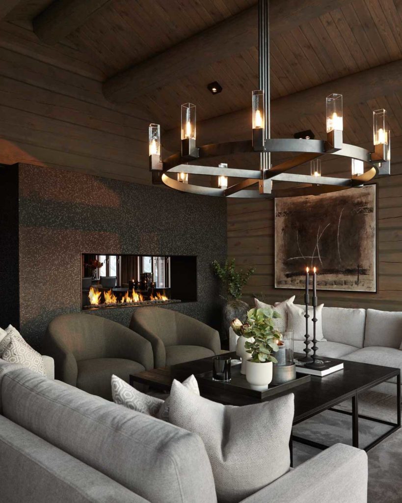 A beautiful living room on a beautiful log cabin. A large chandelier hangs from the ceiling and there is a fire in a minimalist black gas fireplace.