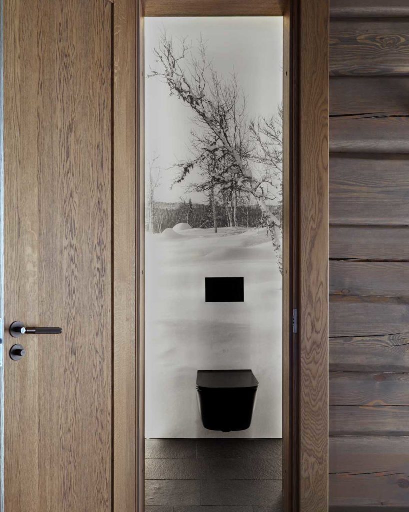 En liten toalett med gråa skifferplattor på golvet och med en bild av vinterklädd natur som tapet bakom en svart toalett. 