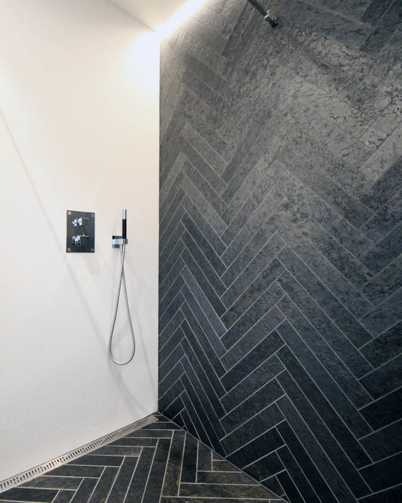 Shower zone with a wall and the floor in black slate tiles laid in a herringbone pattern and a light contrast wall in brushed cement.