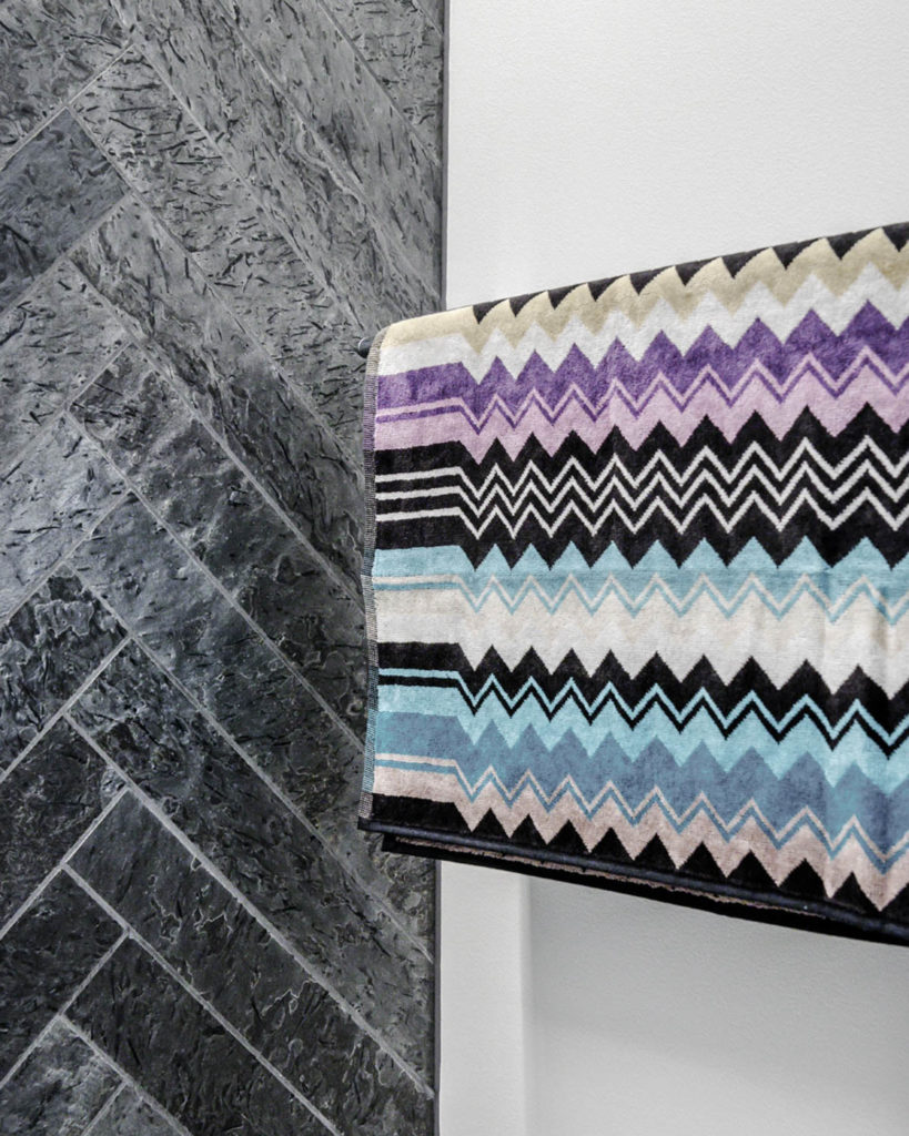 Otta Pillarguri slate tiles in herringbone pattern against a light contrast wall in cement. On the wall hangs a colorful and patterned Missoni towel.