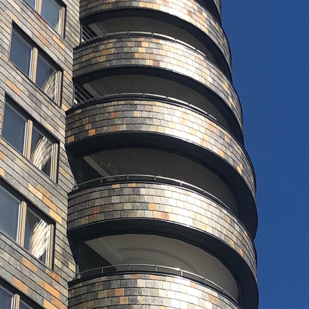 Et høyt leilighetsbygg med runde balkonger kledt med fasadeskifer fra Otta Pillarguri i gylne farger av rust og sort.