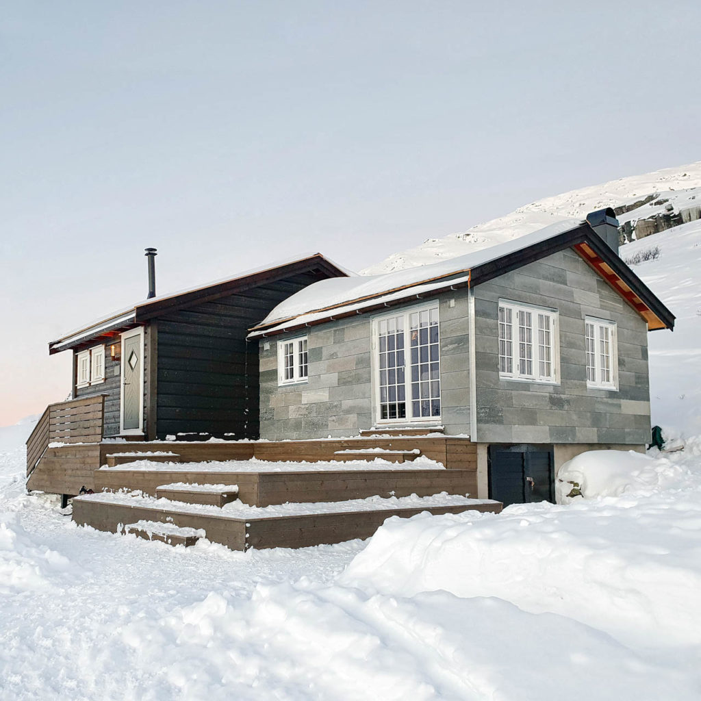 A dark stained mountain cabin with an extension with light gray slate from Oppdal on the facade. There is a lot of snow.