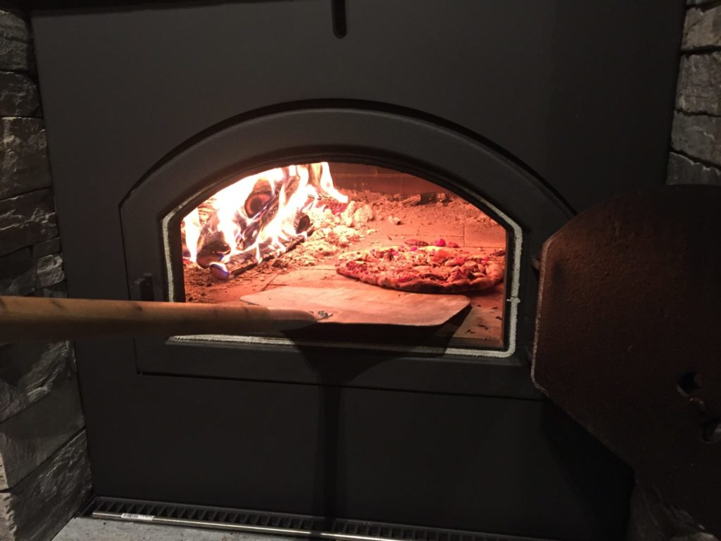 Et nærbilde av en pizza som tas ut med en spade fra en vedfyrt pizzaovn.