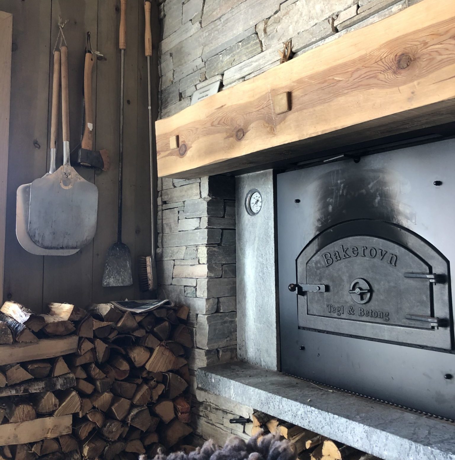 A pizza oven and fireplace bricked with Oppdal quartzite slate. Pizza spatulas and a woodpile stand next to the wall.