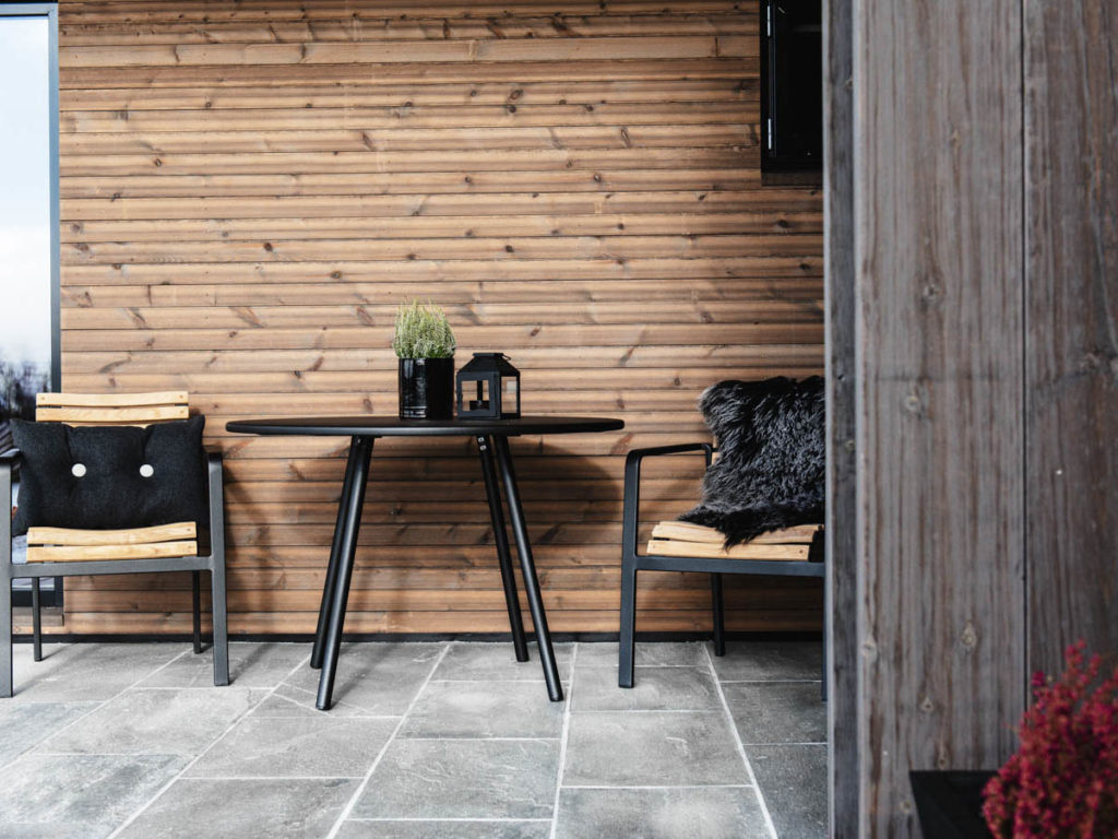 En uteplass med grå skiferflis og smalt brunbeiset trepanel på vegg. Terrassen er møblert med et sort bord og to stoler 
