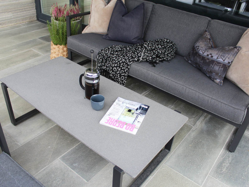 En terrass täckt med skifferplattor. Den är möblerad med en soffgrupp. Det står kaffe på bordet.