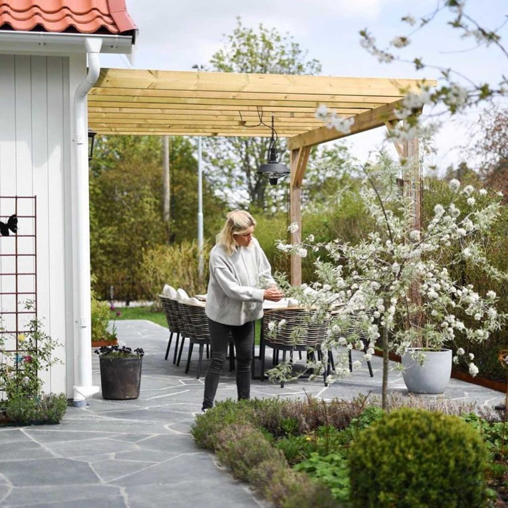 A terrace with pergola and fagstones of Offerdal slate. A lady is tending a beautiful flowering tree.