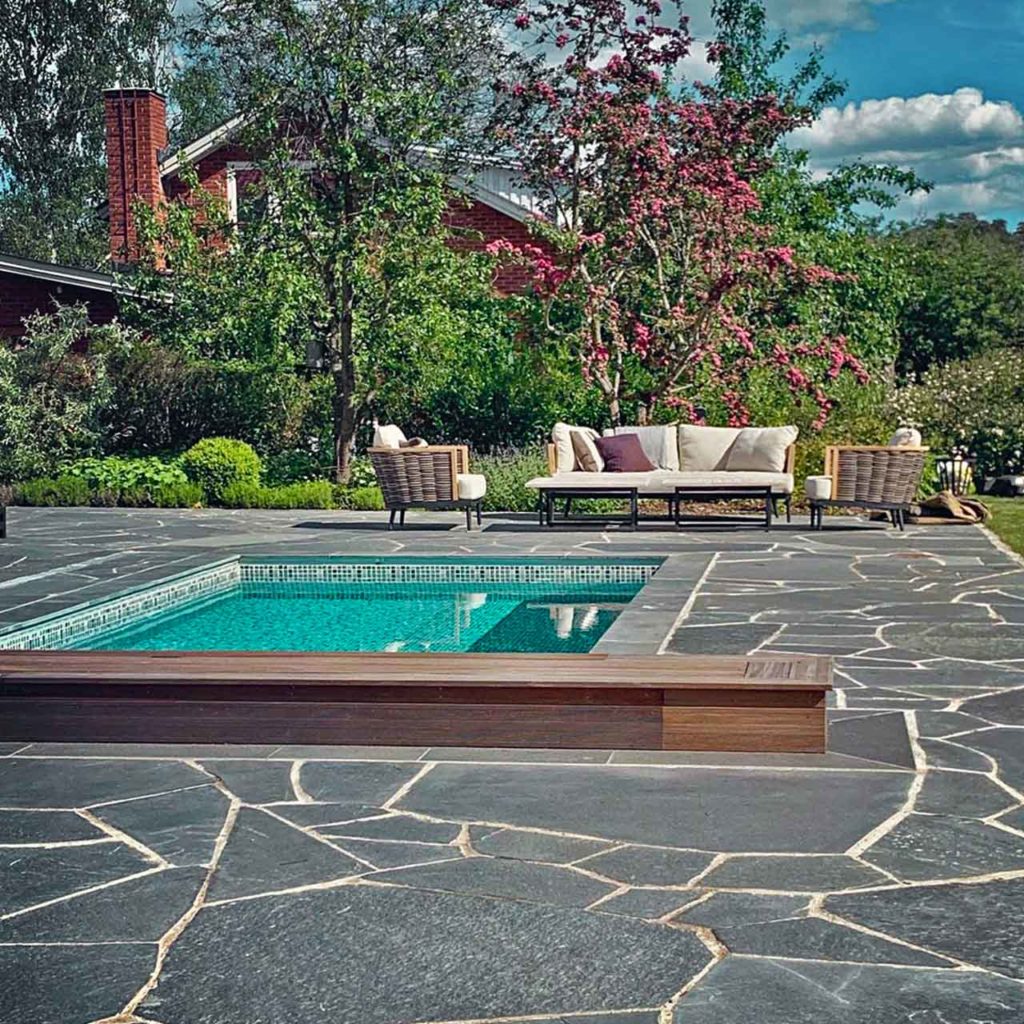 A large paved terrace with swimming pool and seating area. The flagstones are dark gray and there are large flowering trees and shrubs around the terrace.