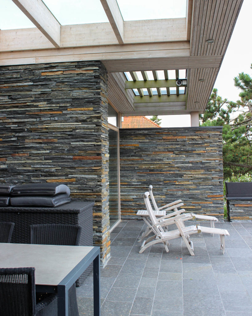 A terrace with walls of brown rusty slate from Otta Pillarguri and with black slate tiles on the ground. On the patio there are sun loungers and a dining area.