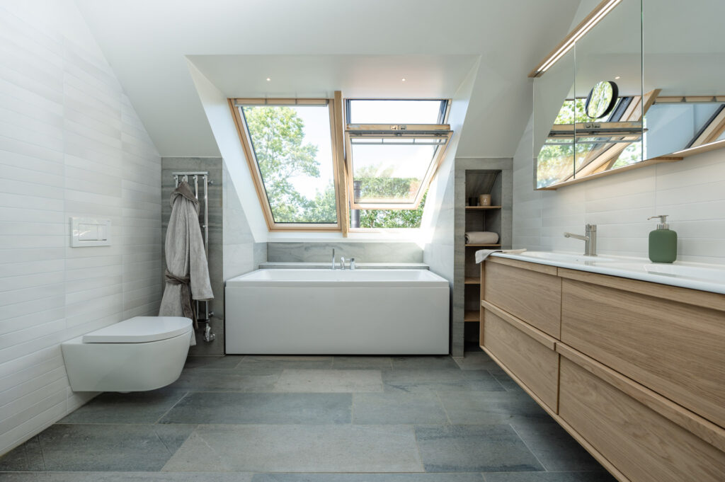 Et bad med lys grå skiferflis på gulv, badekar og badeinnredning i eik
