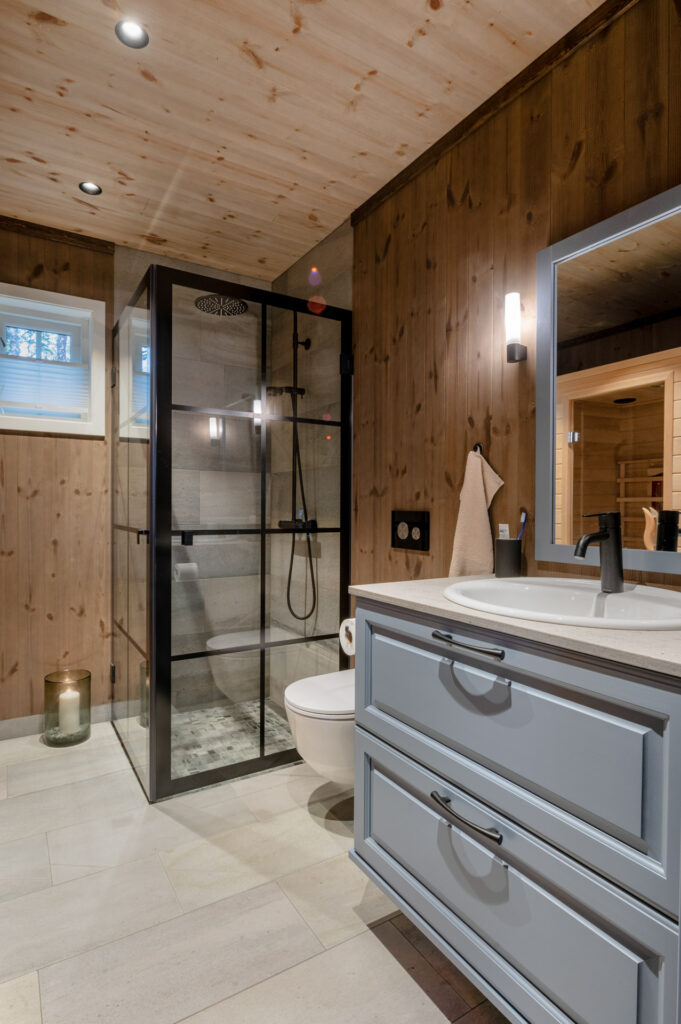 Ett badrum i en stuga med grått kakel och skifferbänkskivor och svart stomme på duschväggarna