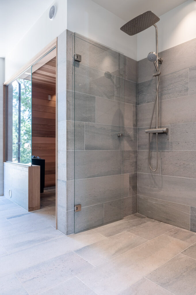 Ett grått badrum med dusch och ingång till bastu, klätt med ljust Oppdalskifferplattor
